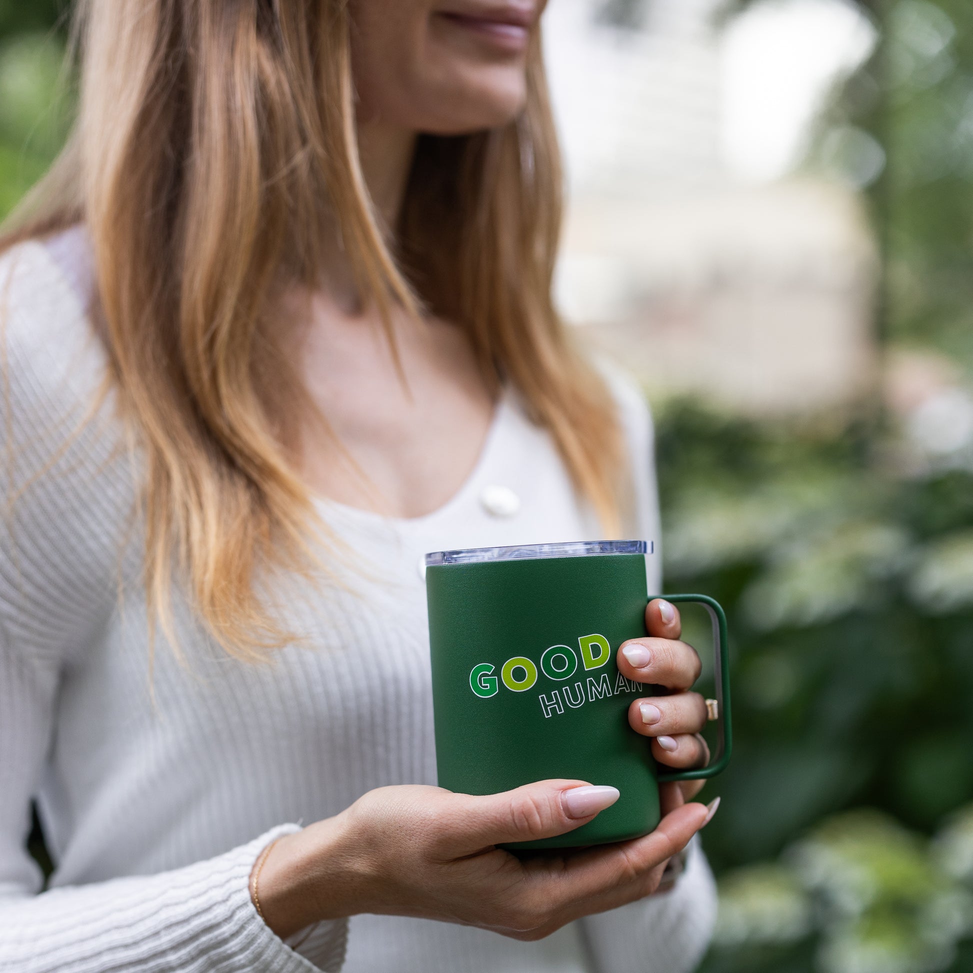 A woman with long hair in a white top holds a green Benebone Good Human Insulated Mug outdoors, with greenery in the background.